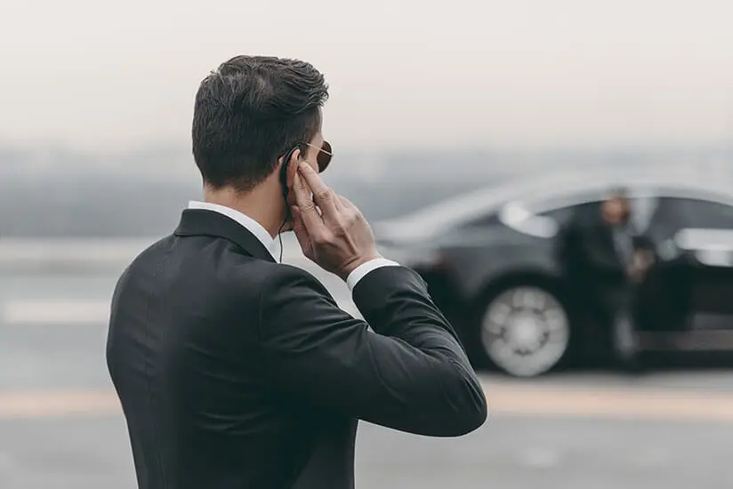 Agent in a dark suit and sunglasses listening to an earpiece beside a parked black car