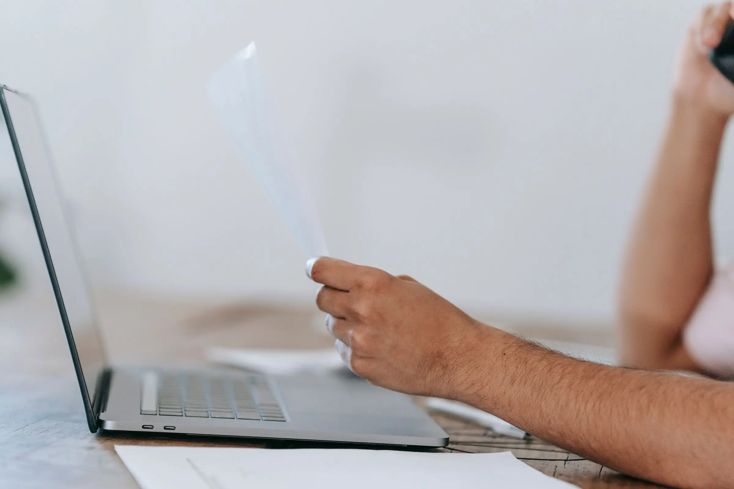 Person on a phone call reading a printed document beside a laptop
